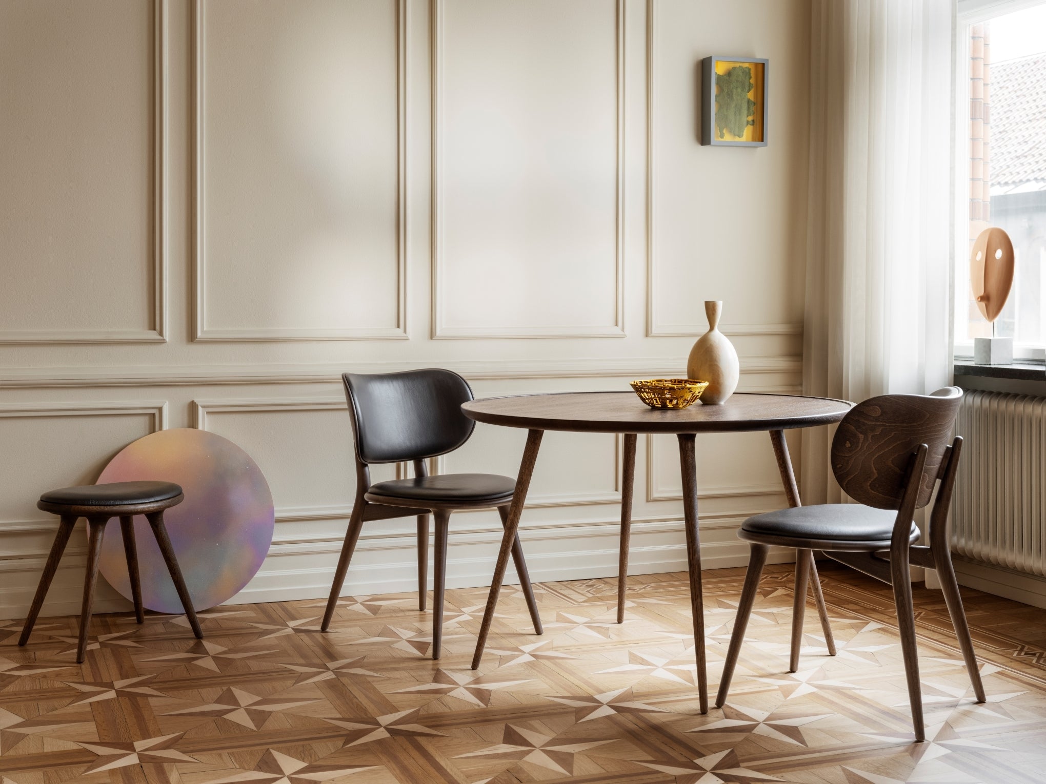 A modern round dining table with brown wooden legs and a multicolored round tabletop, placed in a room with parquet flooring and a decorative wall.
